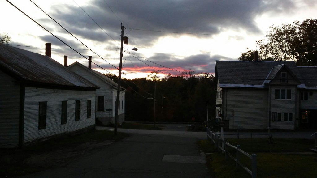 Past old wooden buildings with dingy paint and dark windows, a red-underlit cloud in a pale sky.