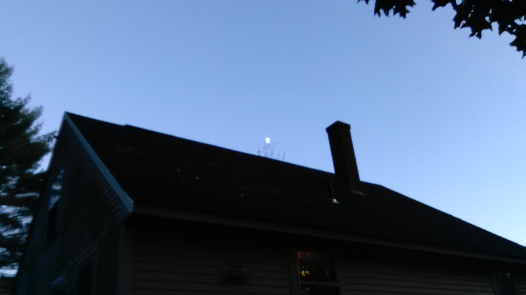 I photographed the upper half of a house, dark against a pale dusk.  A warm glow barely escapes from one window, and the chimney stands starkly against the sky. Pine branches on the left and oak leaves on the upper right, both in silhouette, frame the scene. Wispy bare branches reaching up past the roofline appear to hold the tiny moon aloft.
