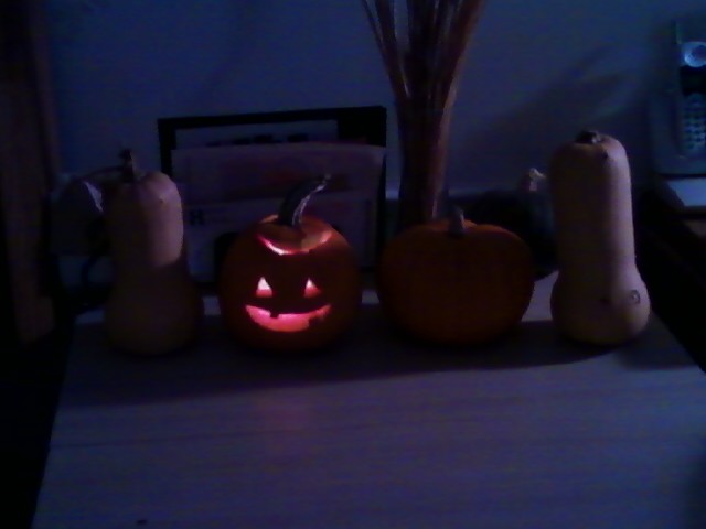 A little lit jack-o'-lantern and a little uncarved pumpkin between 2 butternut squashes on a table in a dim room.